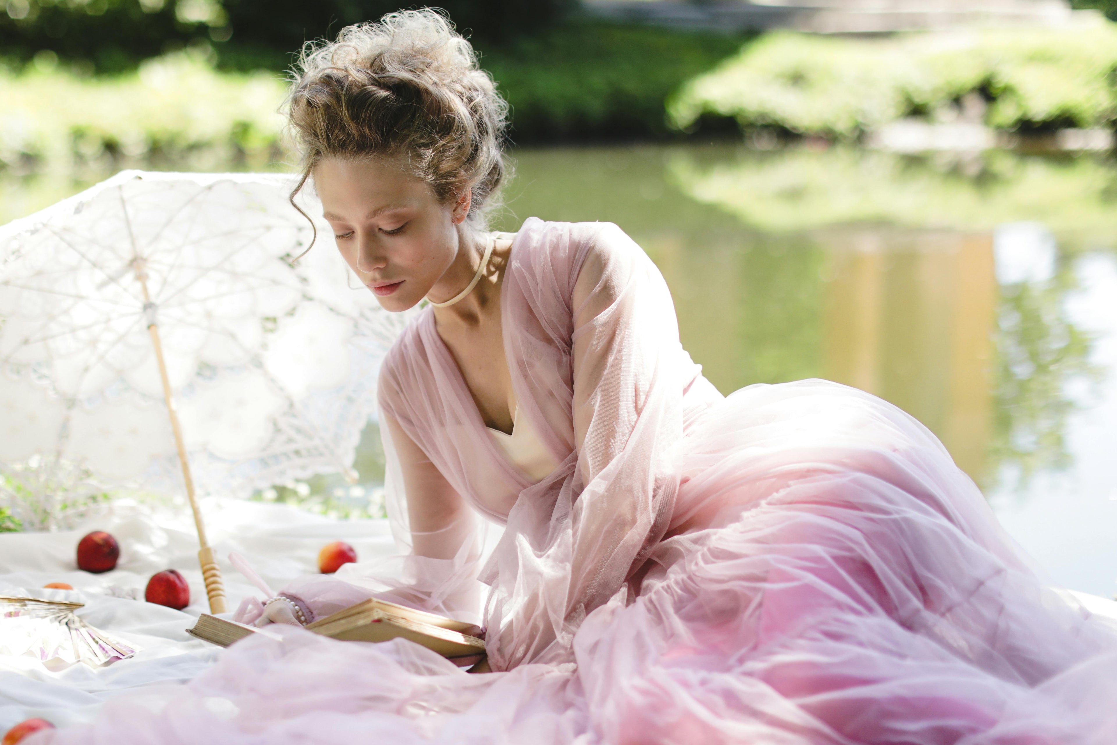 a lady in a pink dress laying on a picnic blanket reading a book with a parasol next to her